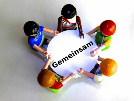 4 Playmobilmännchen von oben gesehen stehen um einen Tisch. Auf dem Tisch steht "Gemeinsam"