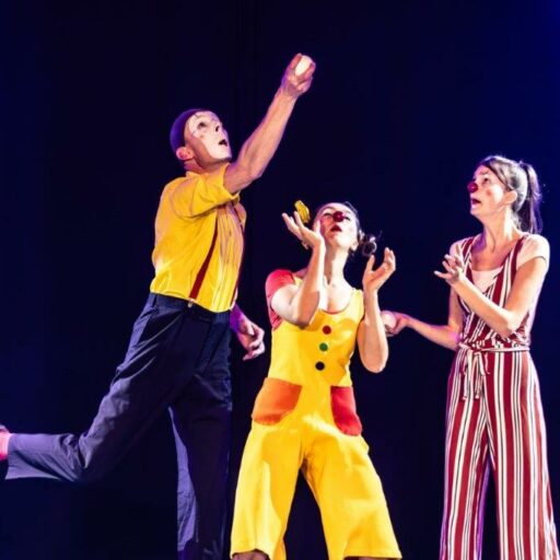 3 Menschen mit Clownsnasen, bunt angezogen auf einer Bühne in Aktion