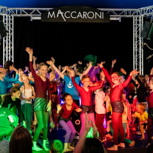 Viele Kinder, winkend auf einer Bühne. Oben hängt ein Schild auf dem steht: "MACCARONI"