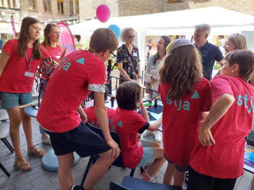 Kinder mit stja-Shirts in Rückenansicht vor einem Tisch mit gebauten fischertechnik-Objekten. Vor dem Tisch stehen Erwachsene, sie sind mit den Kindern im Gespräch