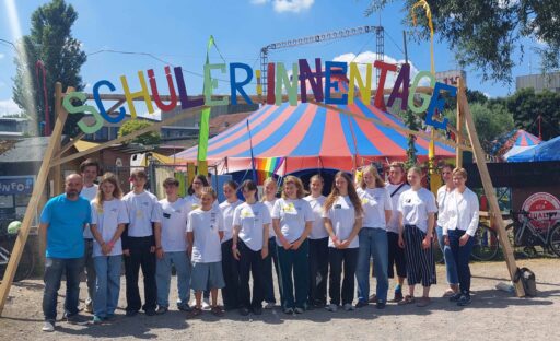 Gruppenbild mit jungen Menschen, Daniel Melchien und einer Frau vor einem Zirkuszelt und einen Gerüst mit dem Schriftzug "Schüler:innentage" in bunten Holzbuchstaben