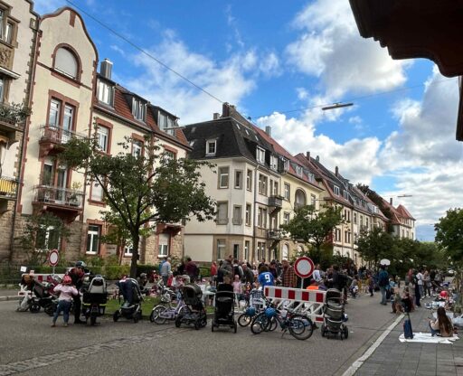 Durch ein Stoppschild und eine Barriere gesperrte Straße. Vor der Absperrung sind kinderwagen und Kinderfahräder abgestellt. Hinter der Absperrung sind Kinder und Eltern zu sehen: spielend oder im Gespräch auf der autofreien gesperrten Straße.