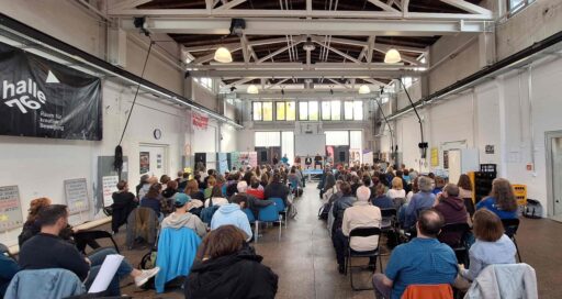 Blick von hinten in eine bestuhlte Halle mit vielen Menschen. Sie blicken auf ein Podium, auf dem 6 Menschen sitzen.