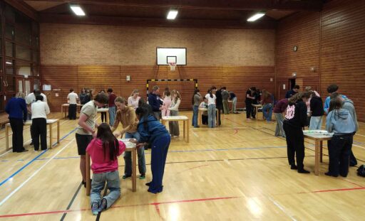 Blick in Sporthalle mit vielen quadratischen Einzeltischen, an denen Jugendliche an der Brezel-Challenge knobeln