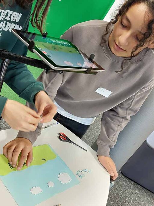 Zwei Jugendliche erstellen einen Film. Über ein auf einem Ständer montiertes iPad werden Fotos gemacht, auf dem Tisch ist ein Blatt mit ausgeschnittenen Wolken und Wiese zu sehen.