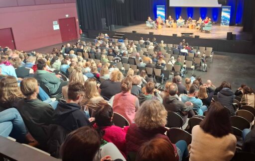 Veranstaltungssaal,die Stuhlreihen sind gut gefüllt mit Blick auf die Bühne. Hier sitzen 8 Menschen im Halbkreis im Gepräch.