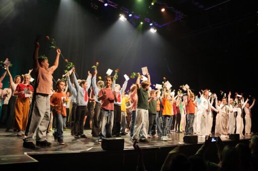 Eine beleuchtete Bühne mit allen jungen Artistinnen und Artisten beim Schlussapplaus, die Arme in die Höhe gereckt, alle haben eine gelbe oder rote Rose in der Hand.