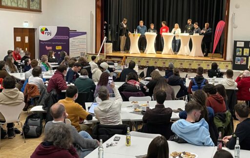 Bühne mit Stehtischen mit 8 Menschen, davor der Raum gefüllt mit Tischen und Menschen auf Stühlen, die auf die Bühne schauen.