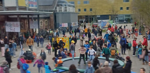 Blick auf den Vorplatz vom Tollhaus mit Erwachsenen und spielenden, tobenden Kindern, im Vordergrund ein großes Trampolin.