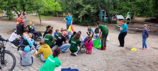 Eine Gruppe von Menschen im Freien an einem Spielplatz. Manche sitzen im Rollstuhl. Auf den Rückansichten von 6 Menschen ist der Aufdruck "Teamer" zu sehen.