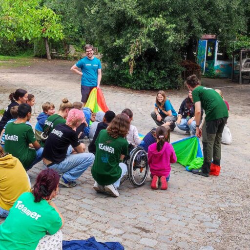 Eine Gruppe von Menschen im Freien an einem Spielplatz. Manche sitzen im Rollstuhl. Auf den Rückansichten von 6 Menschen ist der Aufdruck "Teamer" zu sehen.