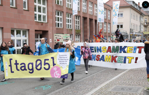 Viele Menschen laufen durch die Stadt, manche kostümiert oder auf Stelzen. Mit Bannern zum Tag 05 mit Texten: "tag05. Europäischer Protesttag zur Gleichstellung von Menschen mit Behinderung", "Gemeinsamkeit statt Einsamkeit".