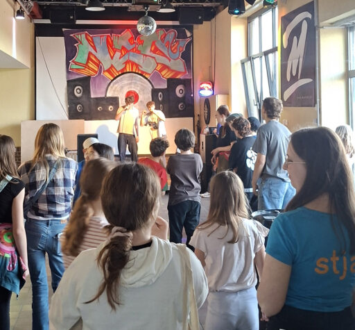 Jugendliche von hinten vor einer Bühne. Auf der Bühne 2 Jugendliche mit Mikrofonen in Aktion.