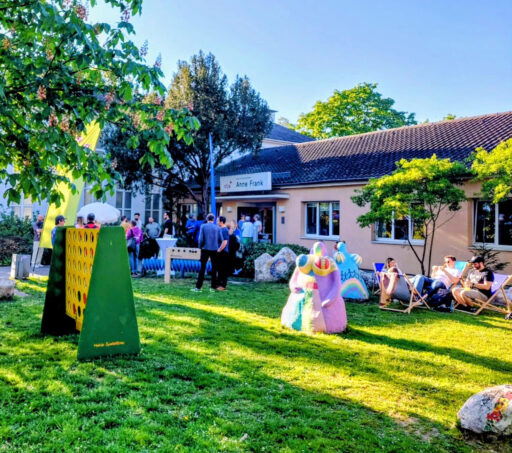 Menschen bei blauem Himmel und Sonnenschein vor dem Anne-Frank-Haus: in Liegestühlen oder stehend im Gespräch. Auf der Wiese auch ein großes 4- Gewinnt-Spiel.