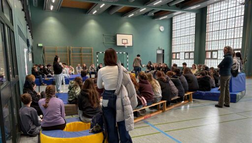 Blick in Sporthalle mit vielen Jugendlichen, die auf Bänken oder Matten sitzen und einer Frau zuhören.