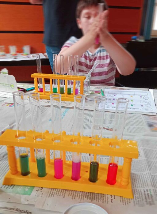 Im Vordergrund Reagenzgläser in einer Stehhalterung mit unterschiedlich farbigen Flüssigkeiten. Im Hintergrund ein Jugendlicher, der ein Reagenzglas zwischen seinen Händen reibt.