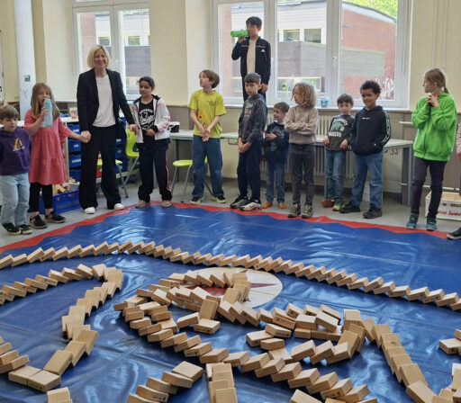 Auf einer blauen Planen liegen Kötze, die gerade per Dominoeffekt umgefallen sind. Drum herum stehen Kinder und eine Erwachsene.