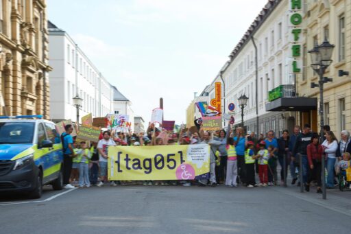 Hinter einem Banner mit der Aufschrift "tag 05 – Europäischer Protesttag zur Gleichstellung von Menschen mit Behinderung" stehen viele Erwachsene und Kinder mit bunten Schildern mit Statements zum tag 05.