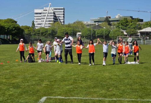 Auf einem Fußballplatz halten sich Mädchen an den Händen und laufen auf die Kamera zu. In der Mitte ihr Trainer Till Hofmann.