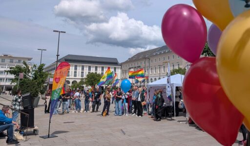Menschen mit Regenbogenflaggen und Luftballons vor dem Karlsruher Rathaus. Im Bilde eine Beachflag vom LA ViE mit dem Schriftzug: Lieb' doch, wen du willst.