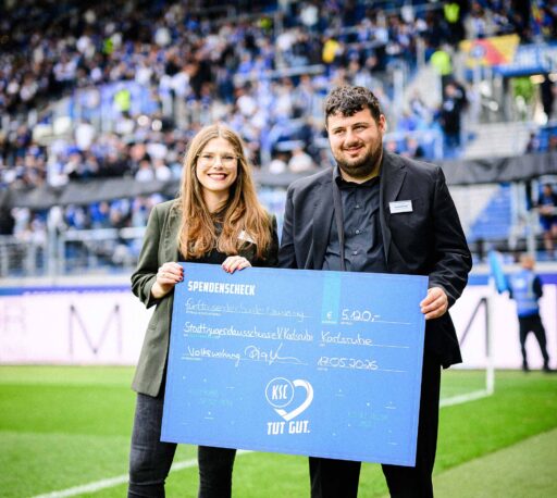 Zwei Menschen halten einen Spendenscheck in Höhe von 5.120 Euro im KSC-Stadion vor gefüllten Rängen