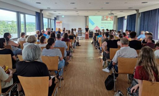 Blick von hinten in einen gut gefüllten Veranstaltungsraum. Zahlreiche Teilnehmende sitzen in Stuhlreihen und schauen zur Referentin sowie zur Präsentationsfolie „Kinderschutz im Verein – Schutzkonzepte leicht gemacht“.
