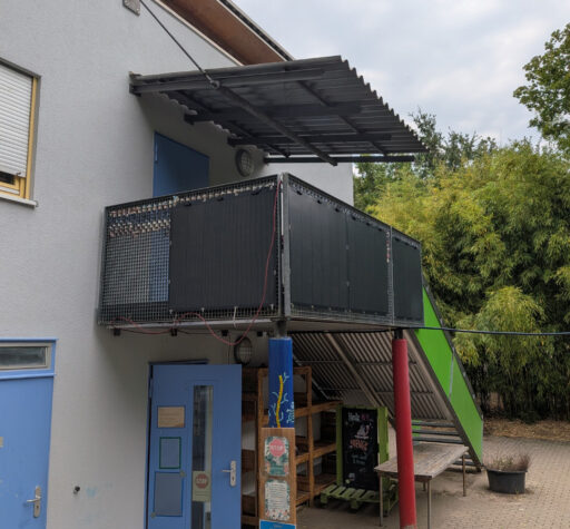 Außenansicht Gebäude mit Treppenaufgang außen zu einem Eingang im ersten Stock mit Balkon. Am Balkon sind außen 4 Solarpannels angebracht.